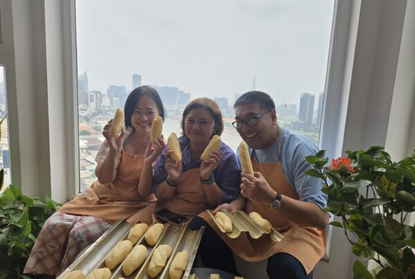Three students hold finished bánh mì loaves at Lua's Kitchen baking class, with the Ho Chi Minh City skyline behind them