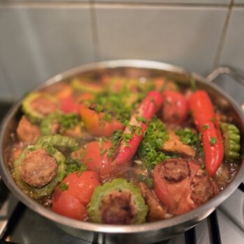 Yong Tau Foo simmering in a stainless pan on a gas hob: stuffed chillies, bitter gourd rings showing the seared pork-and-fish paste, stuffed tomatoes, and scattered spring onion in a clear stock