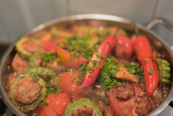 Yong Tau Foo simmering in a stainless pan on a gas hob: stuffed chillies, bitter gourd rings showing the seared pork-and-fish paste, stuffed tomatoes, and scattered spring onion in a clear stock